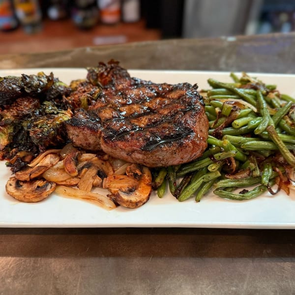 Grilled steak with green beans, roasted Brussels sprouts, mushrooms, and onions on a white plate.